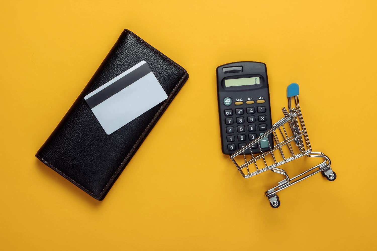 A calculator, wallet, credit card, and micro shopping trolley on a dark yellow background